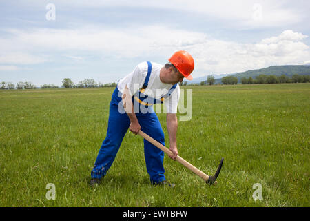 Immagine di un lavoratore in abiti da lavoro e copricapo rigido scavo con un piccone sul verde del campo. Foto Stock