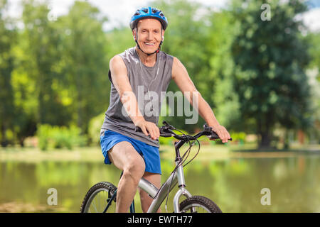 Senior biker con un casco blu in posa sulla sua bicicletta da un laghetto in un parco e guardando la telecamera Foto Stock