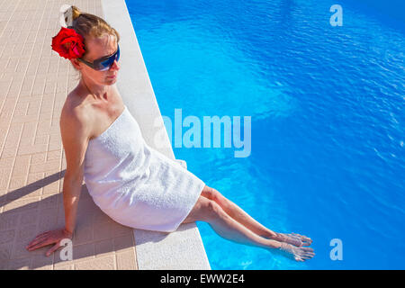 Donna europea che indossa asciugamano bianco rosa rossa e occhiali da sole tenendo le gambe in blu piscina Foto Stock