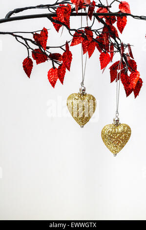 Due golden Natale ornamenti di cuore pendente dal rametto di foglie rosse. Sfondo bianco. Spazio per la copia. Foto Stock