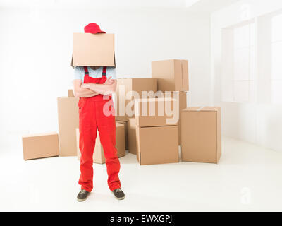 Vista frontale del deliveryman in piedi nella nuova casa con le braccia incrociate contro il petto, la casella che copre la sua testa Foto Stock