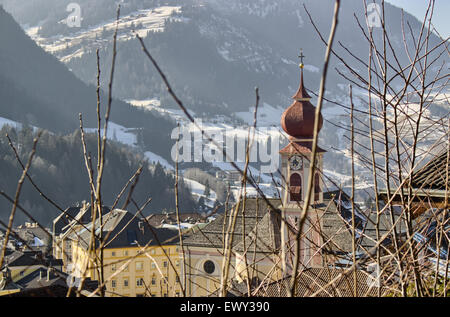 Panorama di Ortisei, villaggio italiano nelle alpi dolomitiche in Italia dietro i rami: case, edifici e la chiesa parrocchiale con campanile tra cime innevate e conifere Foto Stock