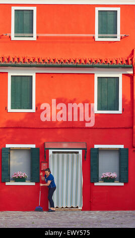 Donna spazzare davanti di colore rosso brillante casa di sunrise Burano Laguna di Venezia Veneto Italia Europa Foto Stock