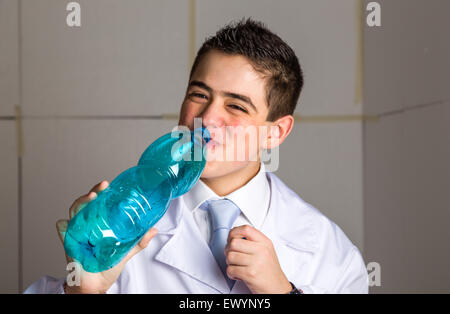 Un ragazzo medico in cravatta blu e mantello bianco di bere acqua da bottiglie di plastica. La sua pelle acne non ha ben ritoccata Foto Stock