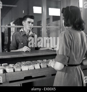 Storico degli anni cinquanta, giovani donne bibliotecario al contatore in una libreria circa per controllare una carta vassoio di indice in cerca di un libro il giovane sta cercando. Foto Stock