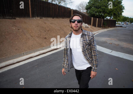 Giovane maschio hipster guardando la fotocamera mentre si camminava sulla strada suburbana Foto Stock