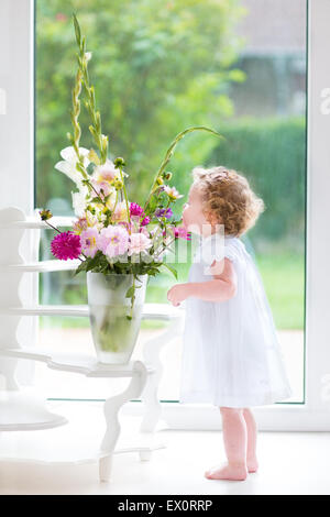 Bambino adorabile ragazza con i capelli ricci che indossa un abito bianco odore di bellissimi fiori accanto a un vento e porta al giardino Foto Stock