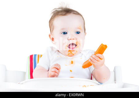 Funny Laughing baby girl mangiando una carota cercando il suo primo solido alimento vegetale seduto in un bianco sedia alta, isolato su bianco Foto Stock