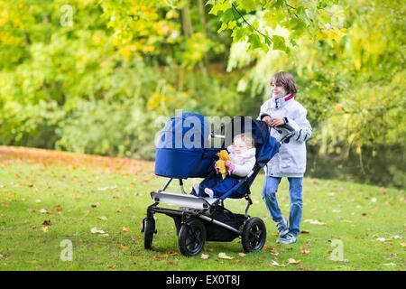 Sorridenti boy a piedi con i suoi piccoli fratellini, un simpatico curly toddler girl e un neonato in un truciolatore doppio passeggino Foto Stock