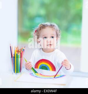 Felice di ridere toddler girl disegno accanto a una finestra sul giardino Foto Stock