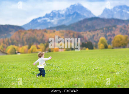 Poco toddler girl in esecuzione in un bellissimo campo tra montagne coperte di neve Foto Stock