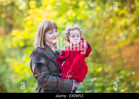 Bellissima ed elegante donna che gioca con un piccolo bambino ragazza in una camicia rossa in un parco d'autunno con colorati alberi giallo Foto Stock