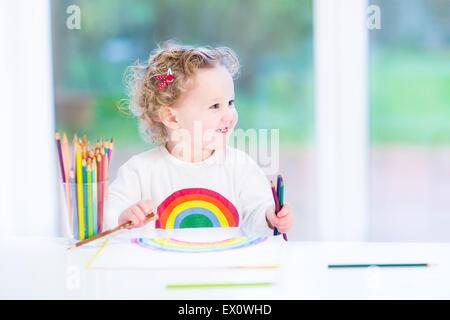 Funny sorridente ragazza toddler disegno un arcobaleno con matite colorate a una scrivania bianca seduto accanto a una grande finestra in giardino Foto Stock