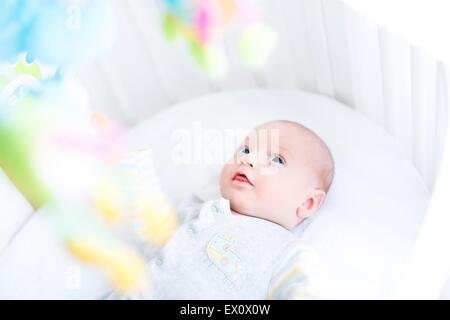 Carino neonato guardando i giocattoli colorati nel suo presepe bianco Foto Stock