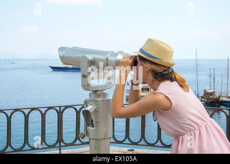 Ragazza giovane guardando attraverso il binocolo pubblica al mare indossando cappello di paglia e abito rosa Foto Stock