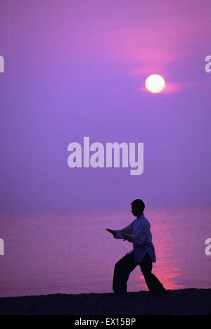 Silhouette di un artista marziale praticare il tai chi al sorgere del sole sulla spiaggia accanto all'oceano. Foto Stock