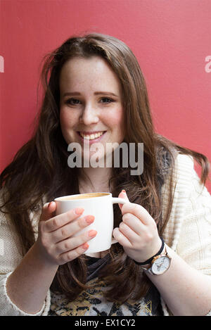 Bella e giovane studente di bere il caffè e sorridente contro la parete rossa. Foto Stock