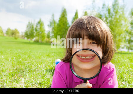 Ragazzo divertente sorrisi attraverso magnifier, fissa sull'erba Foto Stock