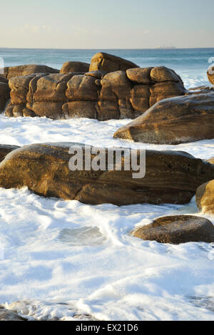 Schiuma bianca da onde turbinano intorno esposti rocce marrone sulla riva del mare Foto Stock