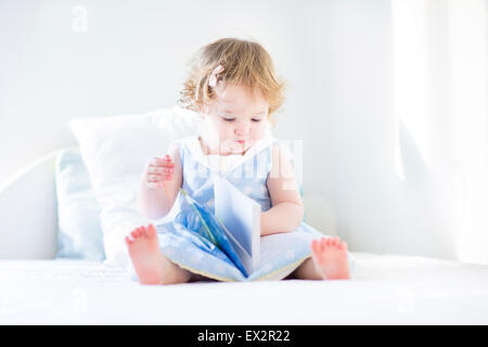 Carino il toddler ragazza con i capelli ricci che indossa un vestito blu seduti su un letto bianco in una luminosa camera da letto la lettura di un libro Foto Stock