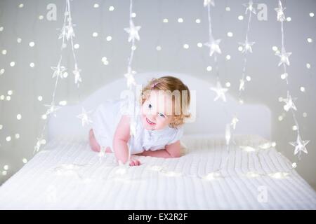 Toddler adorabile ragazza con i capelli ricci che indossa un abito bianco giocando su un letto bianco tra scintillanti luci di Natale Foto Stock