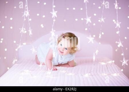 Ritratto di un bimbo adorabile ragazza con bellissimi capelli ricci giocando su un letto bianco tra le belle luci di Natale in rosa Foto Stock