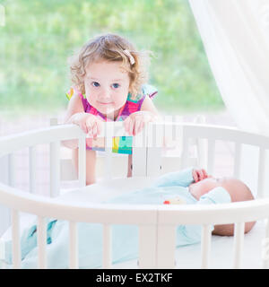 Carino il toddler girl giocando con il suo neonato fratello in un bianco letto rotondo accanto a una grande finestra con vista sul giardino Foto Stock