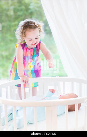 Carino ridere toddler girl giocando con il suo neonato fratello posa in bianco presepe tondo accanto alla grande finestra con vista sul giardino Foto Stock