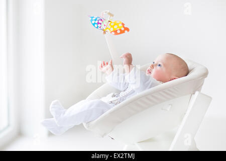 Carino neonato boy guardando un colorato giocattolo mobile seduti in un bianco ad alta sedia accanto a una finestra Foto Stock