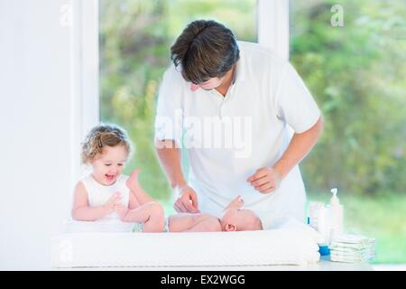 Carino neonato guardando a suo padre e a sua sorella toddler cambiando il pannolino in una stanza con una grande finestra Foto Stock