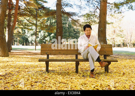 Senior giapponese uomo seduto su una panchina con un libro in un parco della città in autunno Foto Stock