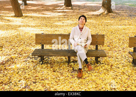 Senior giapponese uomo seduto su una panchina con un libro in un parco della città in autunno Foto Stock