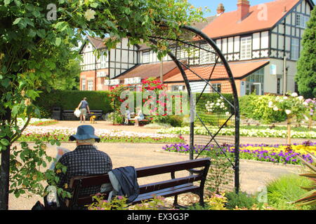 Un uomo si siede su una panchina sotto un arco di rose Hall Leys Park, un tradizionale British park a Matlock, Derbyshire, in Inghilterra, Regno Unito Foto Stock