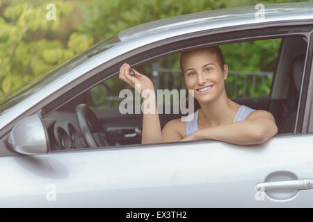 Giovane bella donna seduta dentro la sua auto, sorridente alla telecamera mentre che mostra i tasti sulla sua mano. Foto Stock