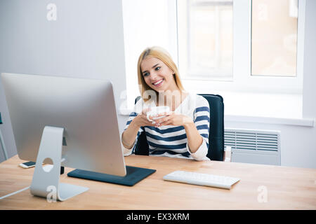 Felice designer femmina di sedere a tavola con i computer desktop e di bere il caffè in ufficio Foto Stock