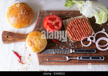 Ingredienti per fare in casa burger sul tagliere di legno, servita con carni coltello e forchetta sulla tovaglia bianca. Ruggine scuro Foto Stock
