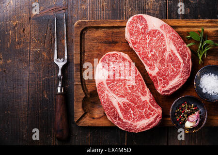 Due materie fresche di carne in marmo nero bistecca angus bistecca, condimenti a base di carne e sulla forcella scuro dello sfondo in legno Foto Stock