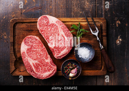 Due materie fresche di carne in marmo nero bistecca angus bistecca, condimenti a base di carne e sulla forcella scuro dello sfondo in legno Foto Stock