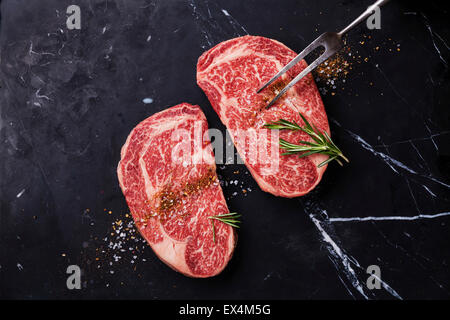 Due materie fresche di carne in marmo nero bistecca angus bistecca, condimenti a base di carne e sulla forcella di marmo scuro dello sfondo Foto Stock