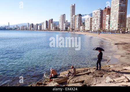 Un uomo ripari dal sole sotto un ombrellone in riva al Mediterraneo a Benidorm, Spagna. Foto Stock