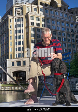 Rilassata uomo maturo con cane seduto fuori sul suo houseboat guardando al suo iPad aria computer tablet Canary Wharf London E14 Foto Stock
