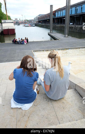 Due giovani donne che si siedono sui gradini accanto all'acqua caratterizzano a Harbourside Bristol, 8 luglio 2015 Foto Stock