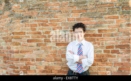 Bel ragazzo caucasico che indossa una camicia bianca e una cravatta regimental con Rosso, Fucsia, arancione, blu indaco e strisce bianche attraversa le braccia davanti ad un muro di mattoni al tramonto Foto Stock