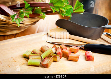 La preparazione di rabarbaro. Di rabarbaro tagliato steli su schede di taglio con coltelli, cucchiai, pala, cesto con gambi freschi in background Foto Stock