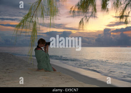 Una giovane donna siede sulle rive sabbiose dell'Oceano Indiano e fotografare il tramonto, Denis Island, Seicelle Foto Stock