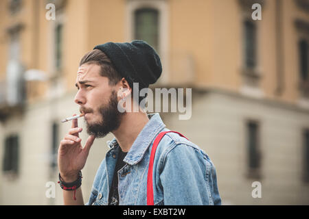 Giovane bello barbuto hipster uomo fumatori di sigarette in città Foto Stock