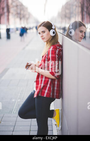 Giovane bella bionda hipster donna nella città con ukulele e cuffie Foto Stock