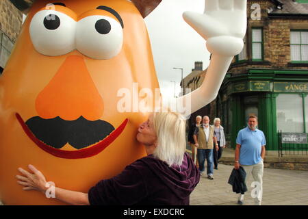 Belper, Derbyshire, Regno Unito. 12 Luglio, 2015. Spud ventola, Freda Raphael piante un bacio su un gigante Mr Potato Head che ha ri-apparve in Derbyshire città. La 7ft-alto statua in fibra di vetro è stato regalato a Belper nel 2001 dalla sua città gemelle, Pawtucket, Rhode Island, Stati Uniti d'America. Dopo essere stato ribattezzato "onstrosity" da parte di alcuni locali, il carattere divisivo realizzati titoli nazionali. È stato soggetto ad atti vandalici e bandito. Ora il spud è stata spruced fino da un gruppo locale di giovani e svelato a Belper's Food festival di oggi. Credito: Deborah Vernon/Alamy Live News Foto Stock