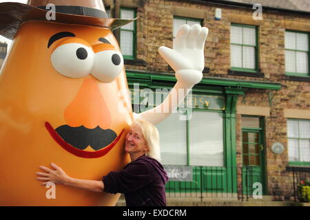 Belper, Derbyshire, Regno Unito. 12 Luglio, 2015. Spud ventola, Freda Raphael abbracci un gigante Mr Potato Head che ha ri-apparve in Derbyshire città. La 7ft-alto statua in fibra di vetro è stato regalato a Belper nel 2001 dalla sua città gemelle, Pawtucket, Rhode Island, Stati Uniti d'America. Dopo essere stato ribattezzato "onstrosity" da parte di alcuni locali, il carattere divisivo realizzati titoli nazionali. È stato soggetto ad atti vandalici e bandito. Ora il spud è stata spruced fino da un gruppo locale di giovani e svelato a Belper's Food festival di oggi. Credito: Deborah Vernon/Alamy Live News Foto Stock