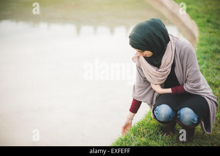 Giovane donna indossa hijab accovacciato accanto al parco lago Foto Stock